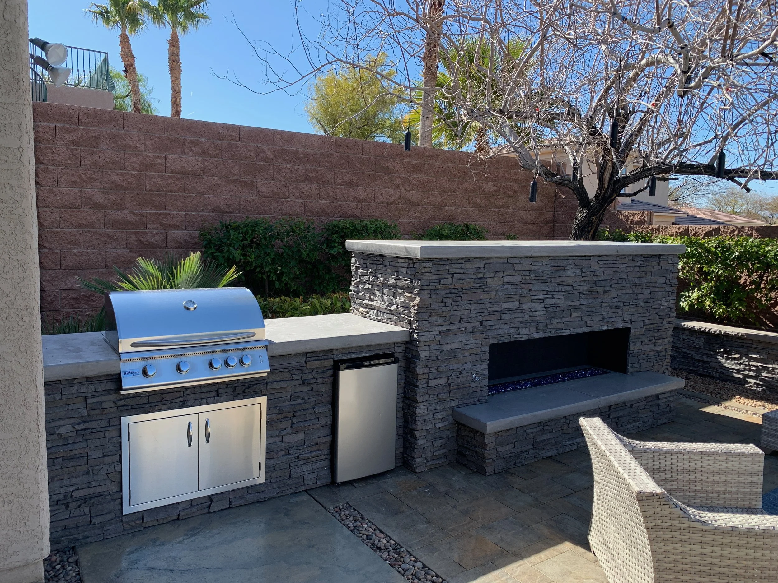 Outdoor kitchen with grill and fireplace beside patio seating in a backyard.