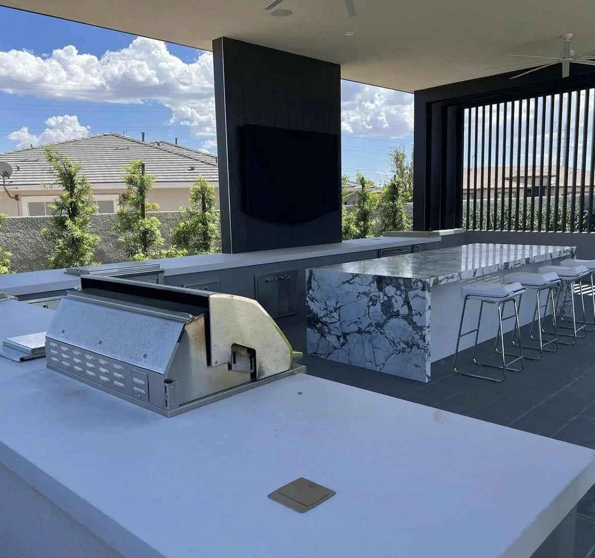 Modern outdoor kitchen with marble countertop, bar stools, built-in grill, and open patio view.