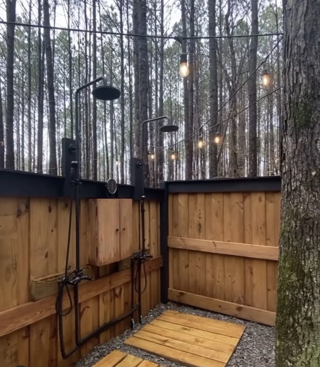 Outdoor shower area with wooden fencing and string lights among trees.