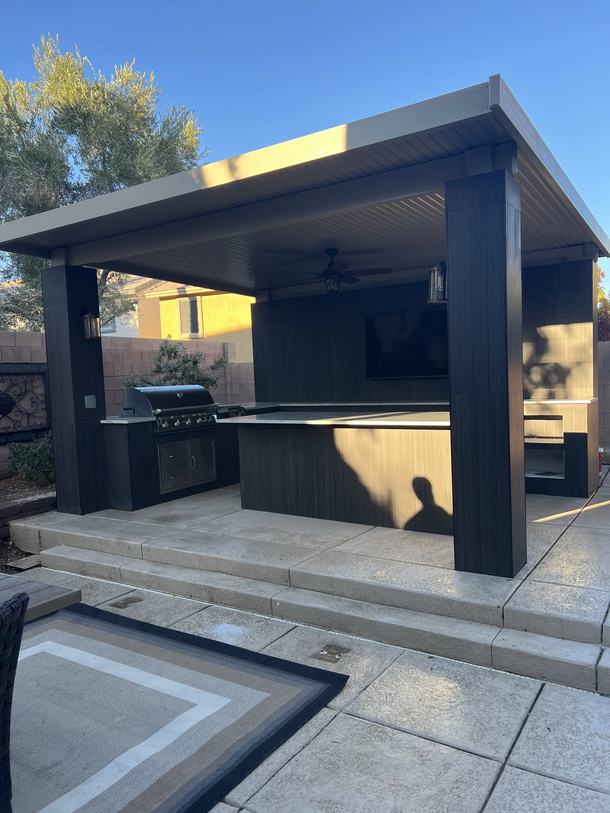 Outdoor kitchen with grill and bar under a pavilion with ceiling fan and lights, adjacent to a patio area with rug.