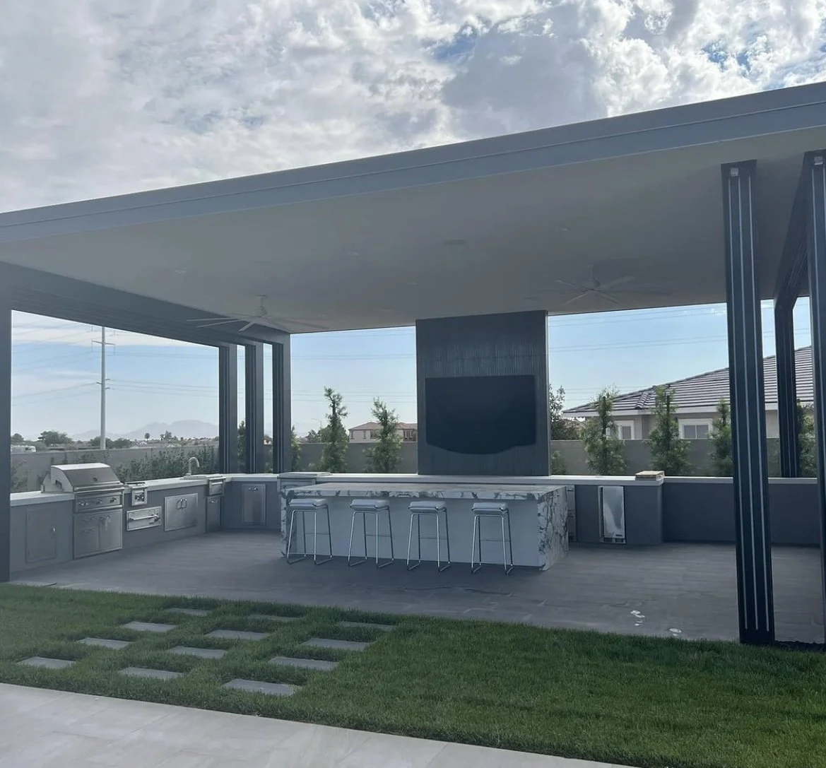 Modern outdoor kitchen with a roofed structure, featuring a grill, countertops, barstools, and a flat-screen TV, surrounded by greenery and a well-maintained lawn.