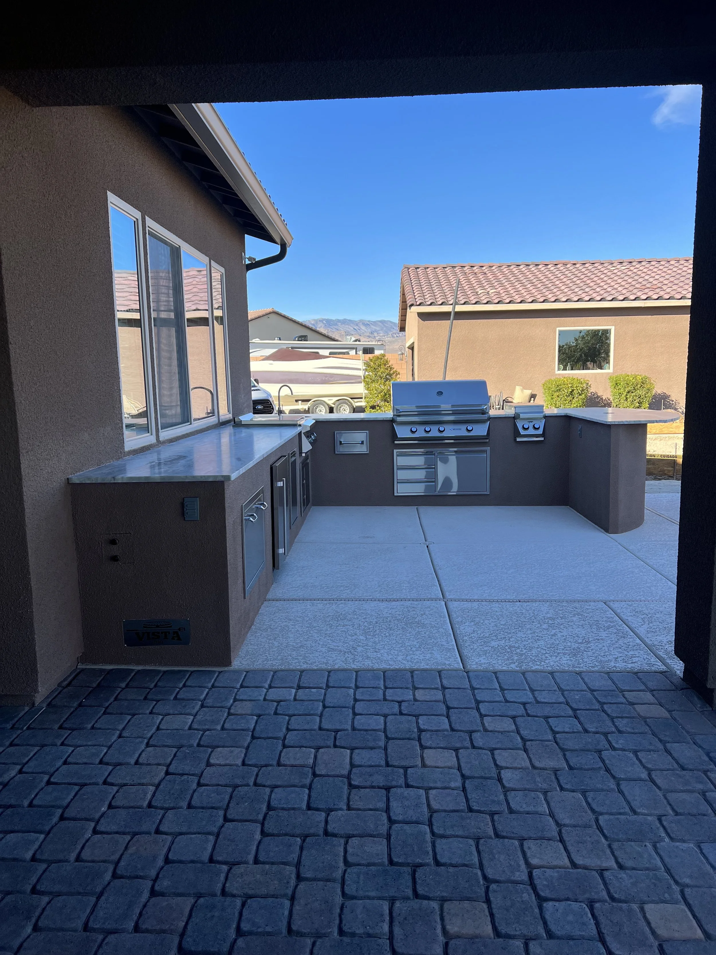 Outdoor kitchen with built-in grill and appliances on a patio with a view of houses and mountains.
