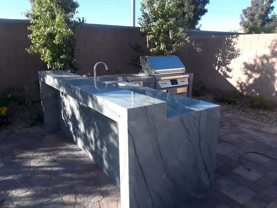 Outdoor kitchen with marble countertop, sink, and grill in a backyard setting with stone pavers and plants.