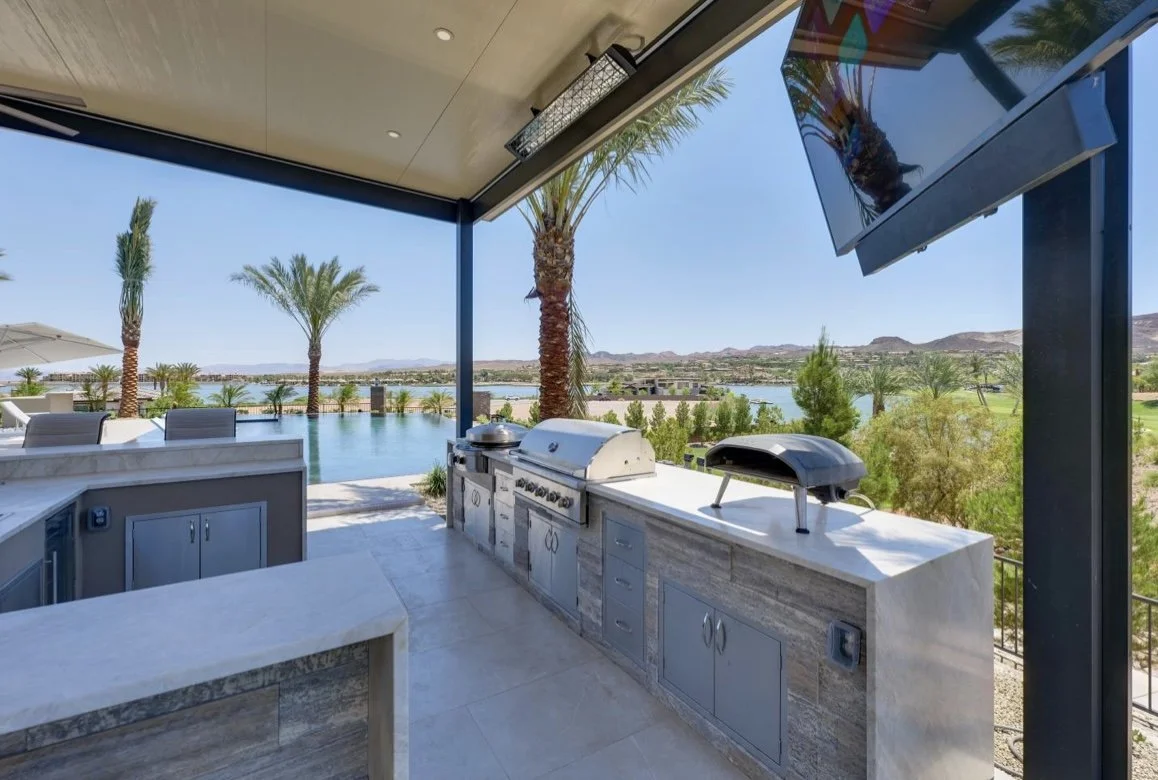 Outdoor kitchen with gas grill and pizza oven overlooking a pool and palm trees with a lake view.