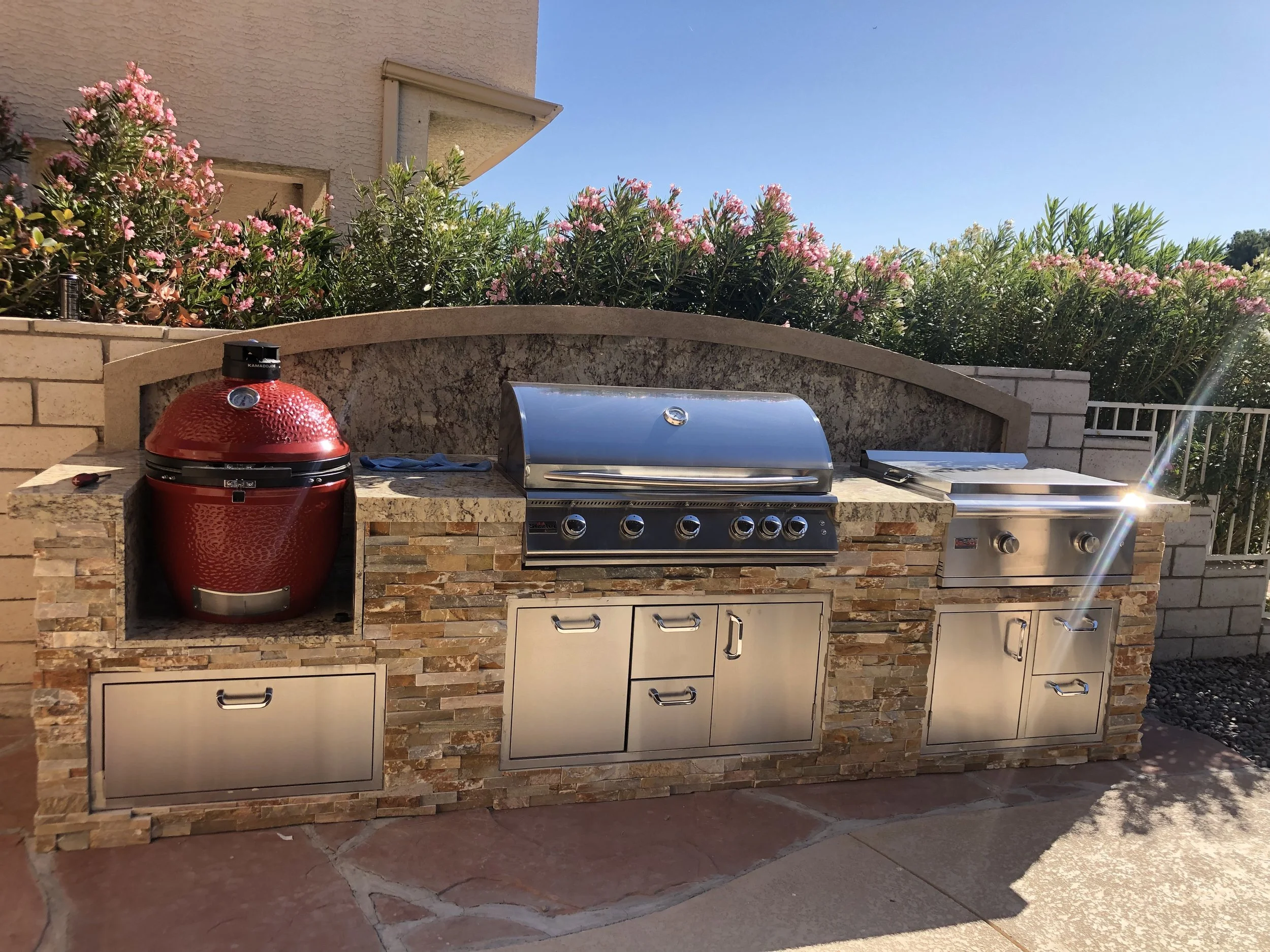 Outdoor kitchen with a red ceramic grill, stainless steel gas grill, griddle, and storage cabinets, surrounded by stonework, with blooming bushes in the background.