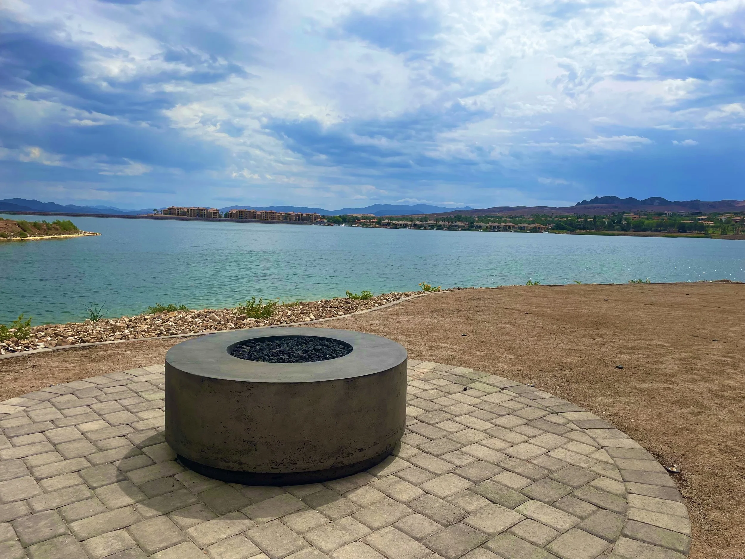 Circular stone fire pit on a paved patio overlooking a lake with mountains in the background, under a partly cloudy sky.