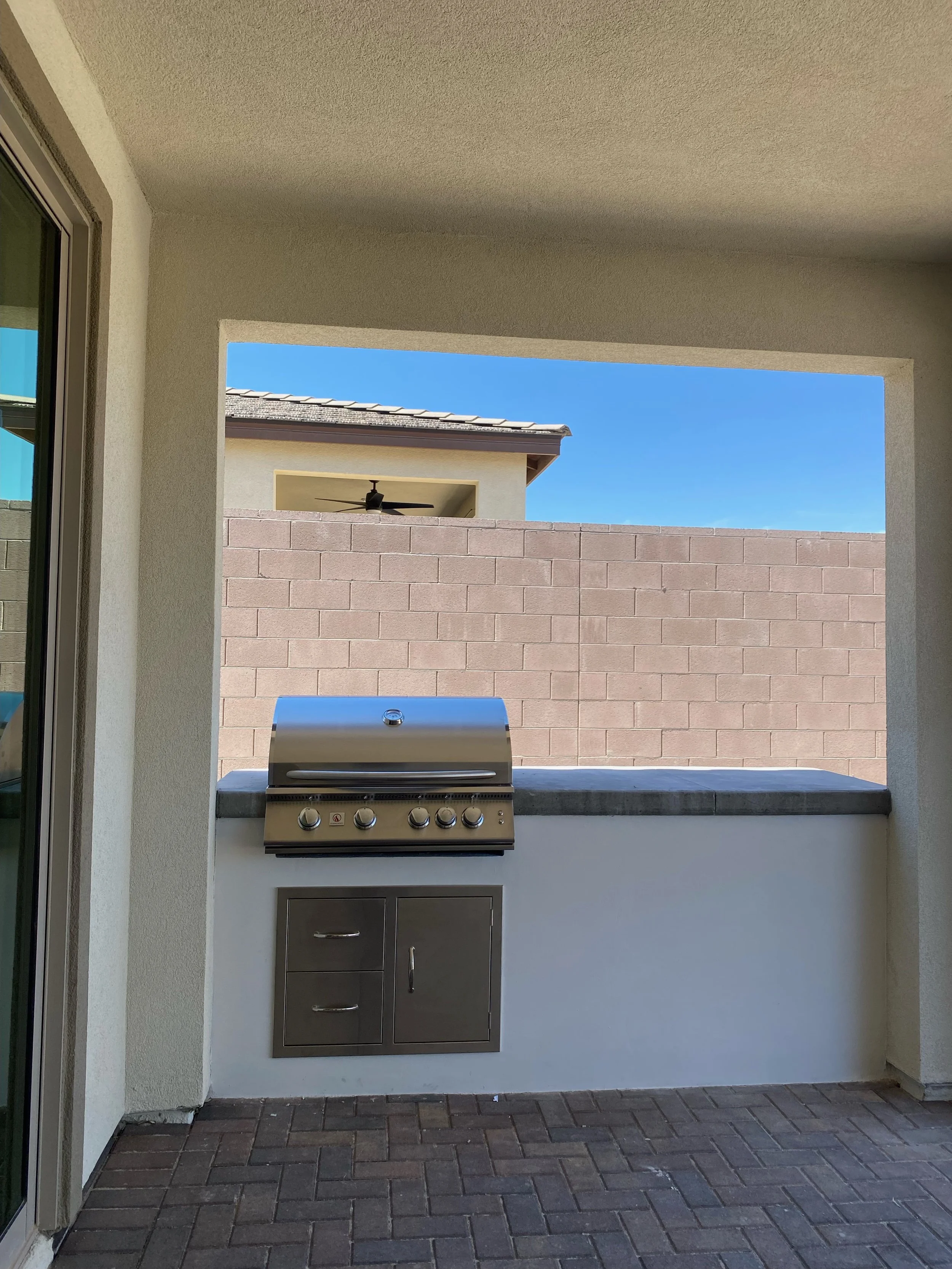 Outdoor patio area with a built-in stainless steel grill, brick flooring, and a partial view of a brick wall and neighboring house.