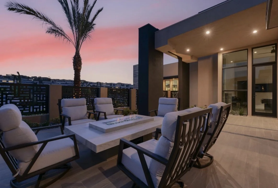 Outdoor patio with cushioned chairs around a fire pit table, modern architecture, and palm tree at sunset.