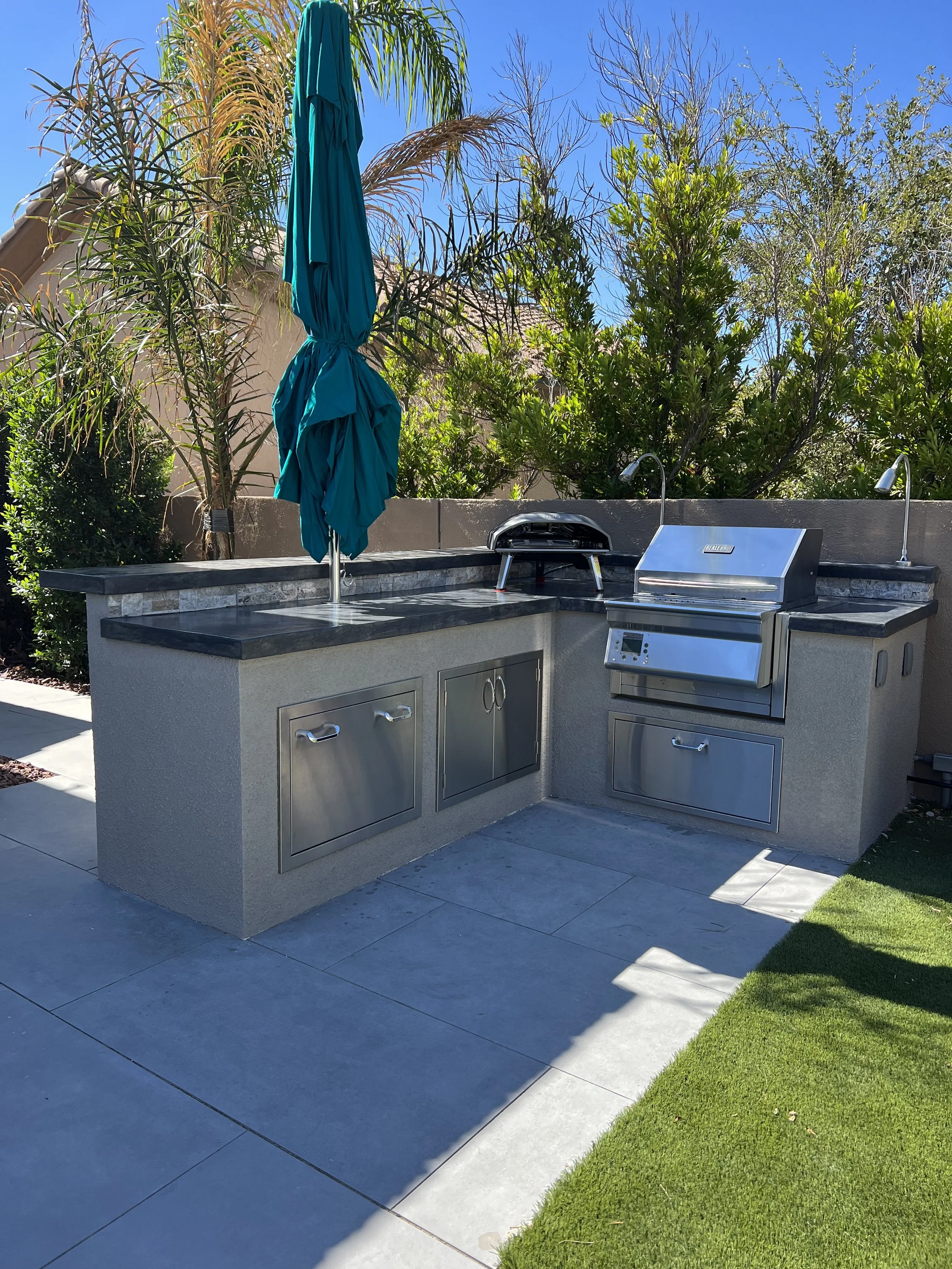 Outdoor kitchen with stainless steel grill and countertop, surrounded by palm trees and greenery.