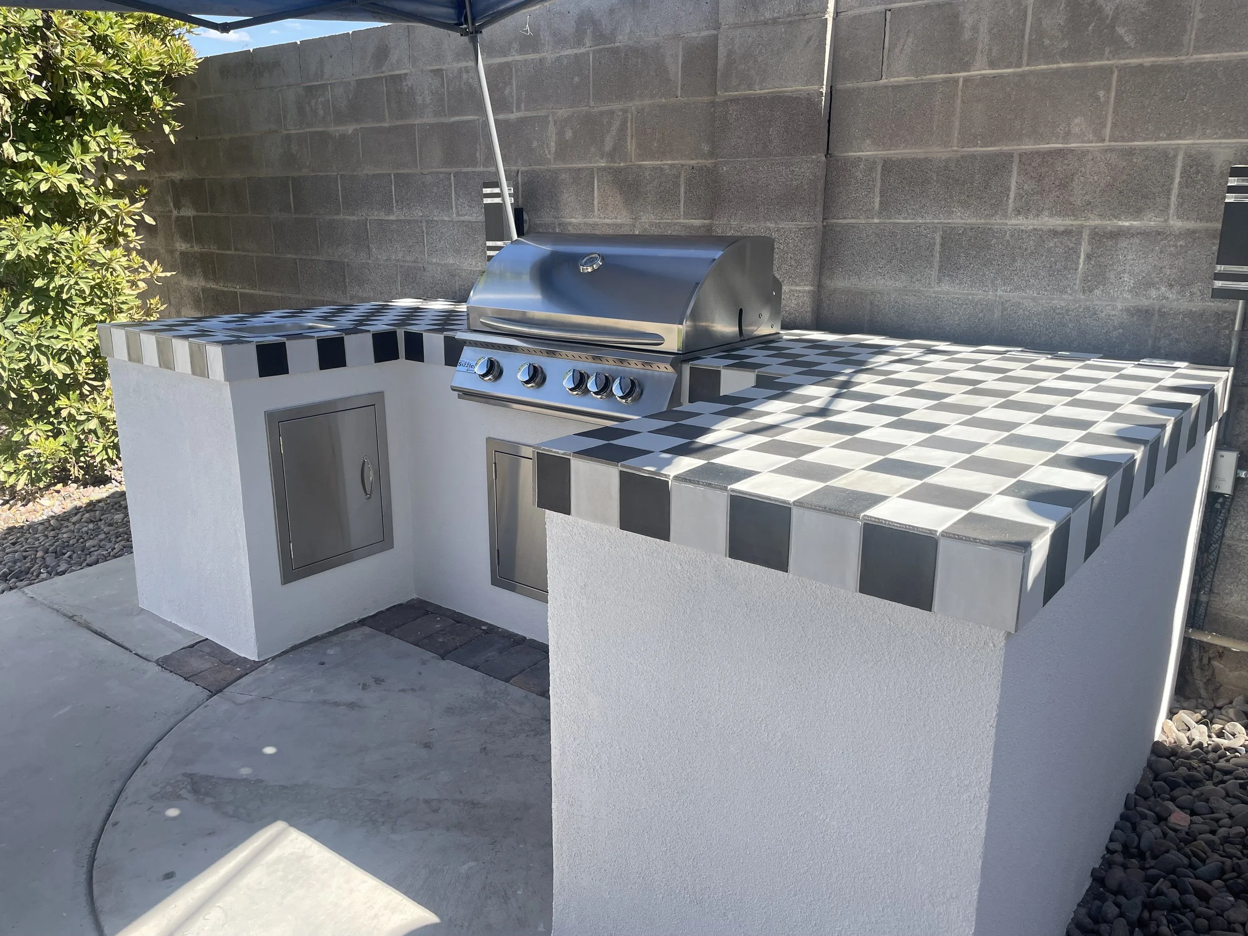 Outdoor kitchen area with a built-in stainless steel grill and checkered tile countertops, set against a block wall.