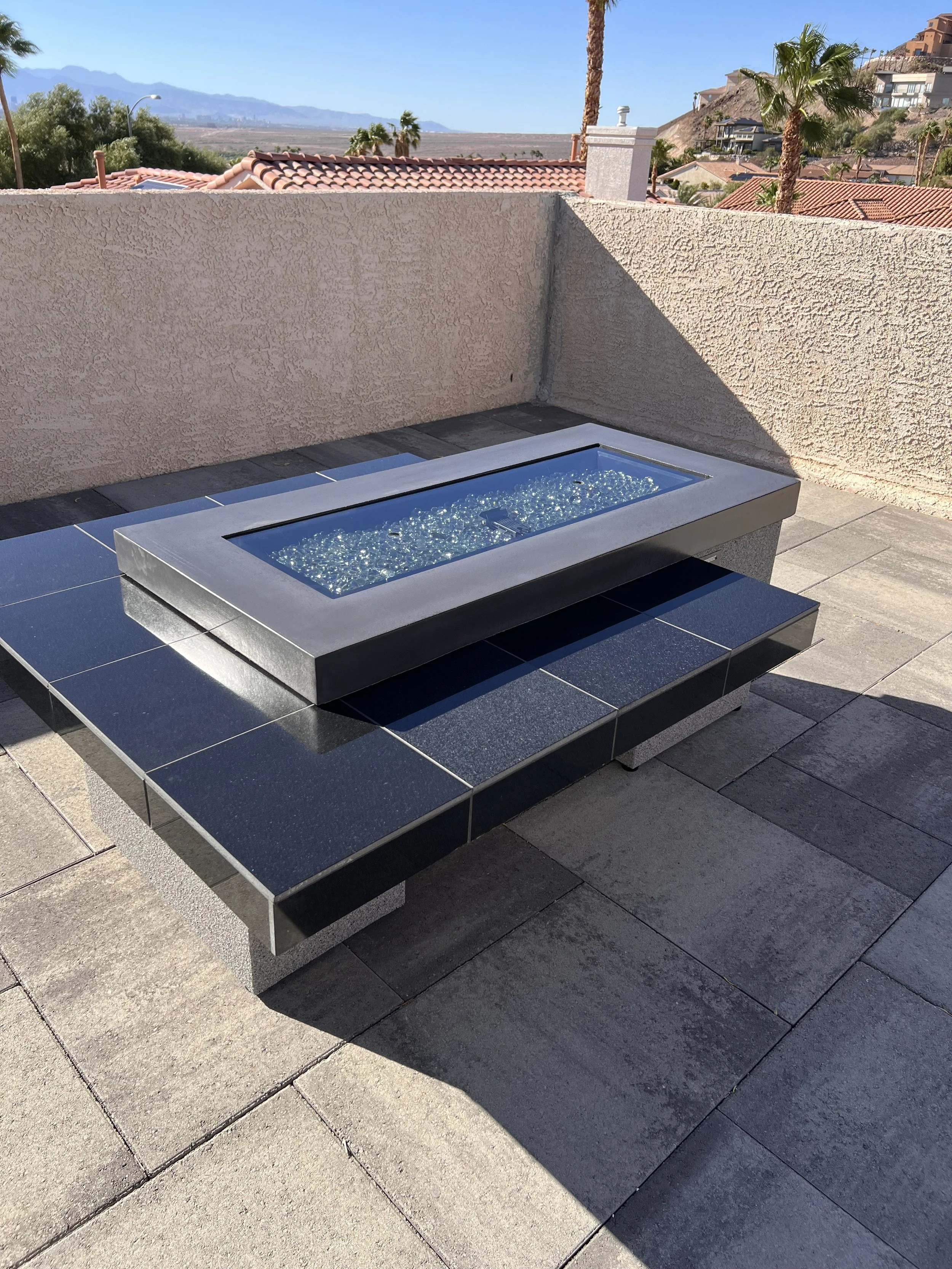 Modern outdoor fire pit with blue glass stones on a patio, surrounded by beige stucco walls and a view of hills and palm trees in the background.