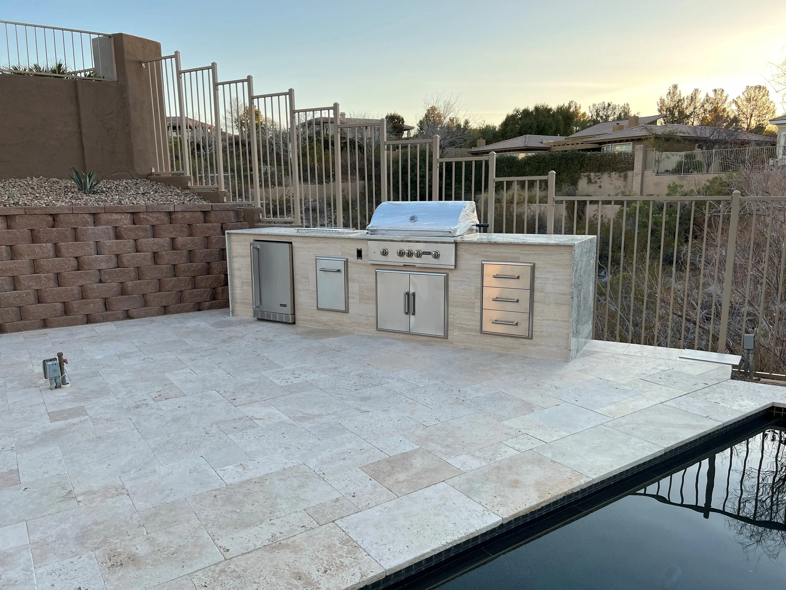 Outdoor kitchen with built-in grill, refrigerator, and drawers on a tiled patio next to a swimming pool, surrounded by metal fencing.