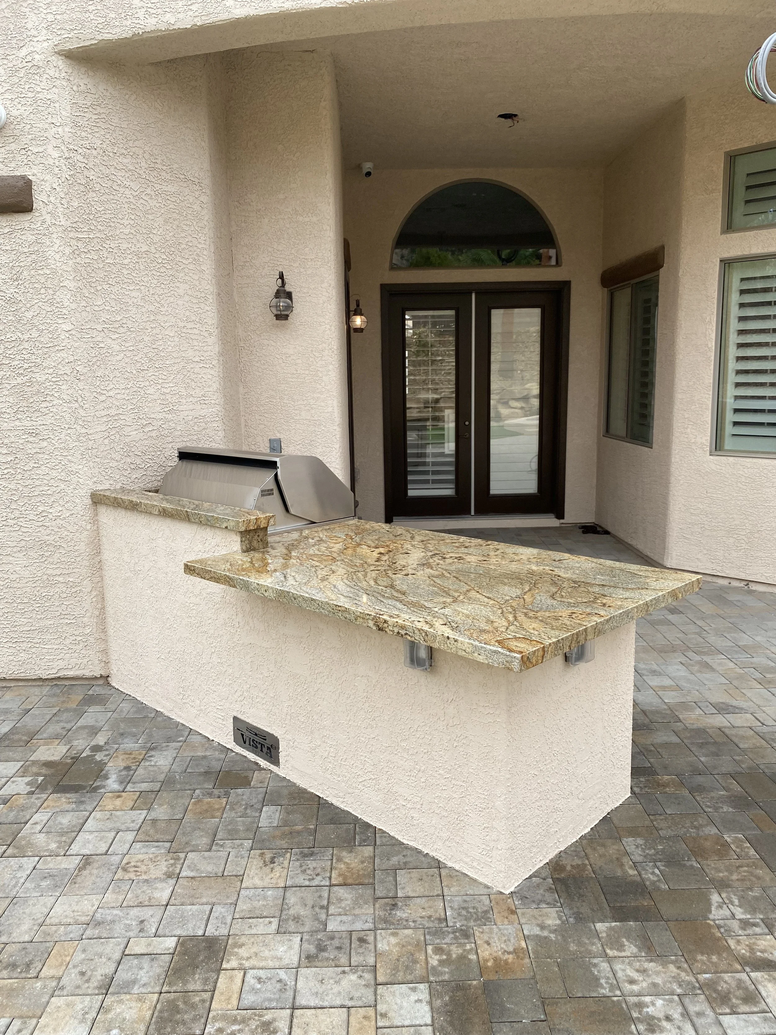 Outdoor kitchen area with a marble countertop and built-in grill, situated on a stone patio with double doors in the background.