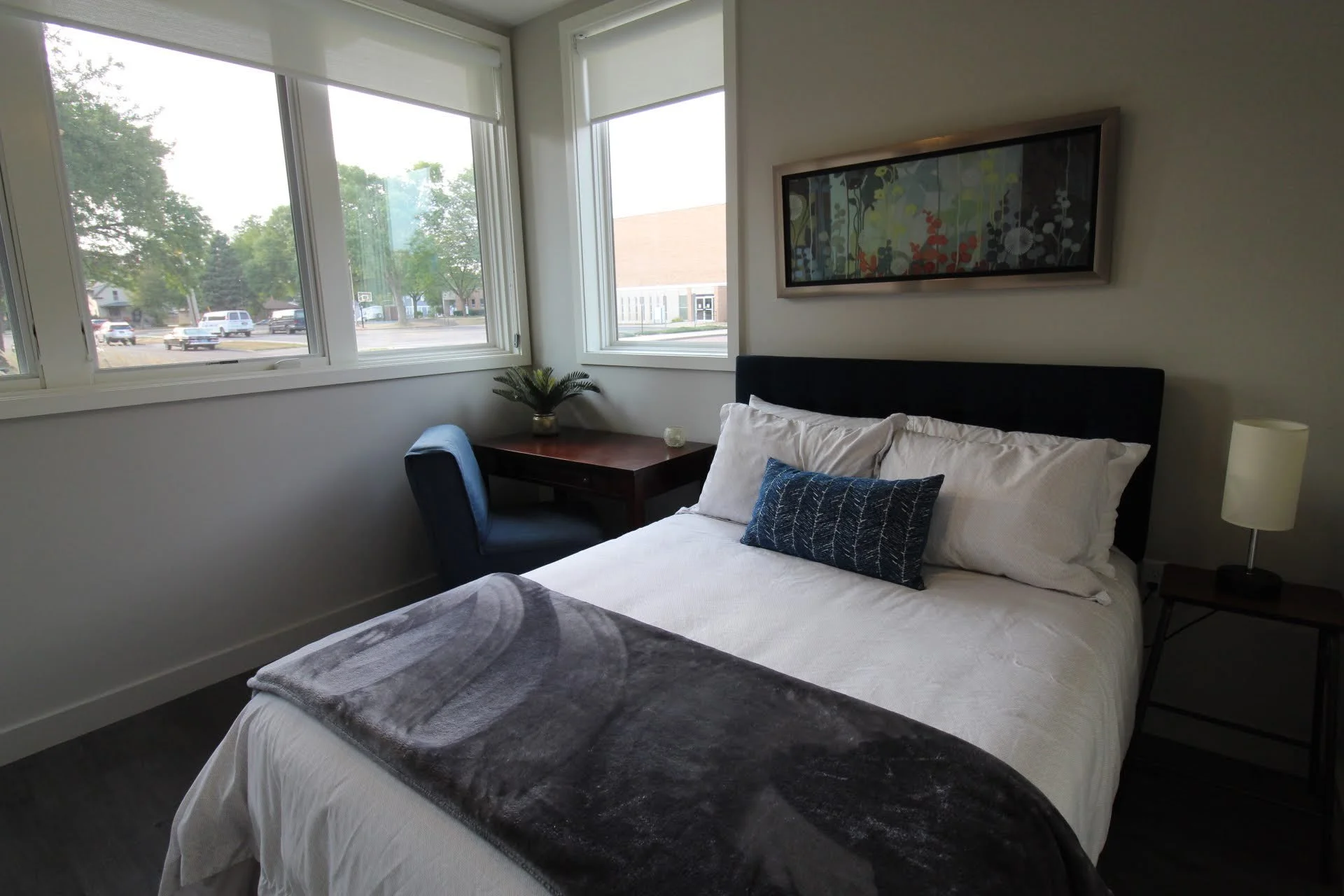 A room with full bed, desk and large windows.