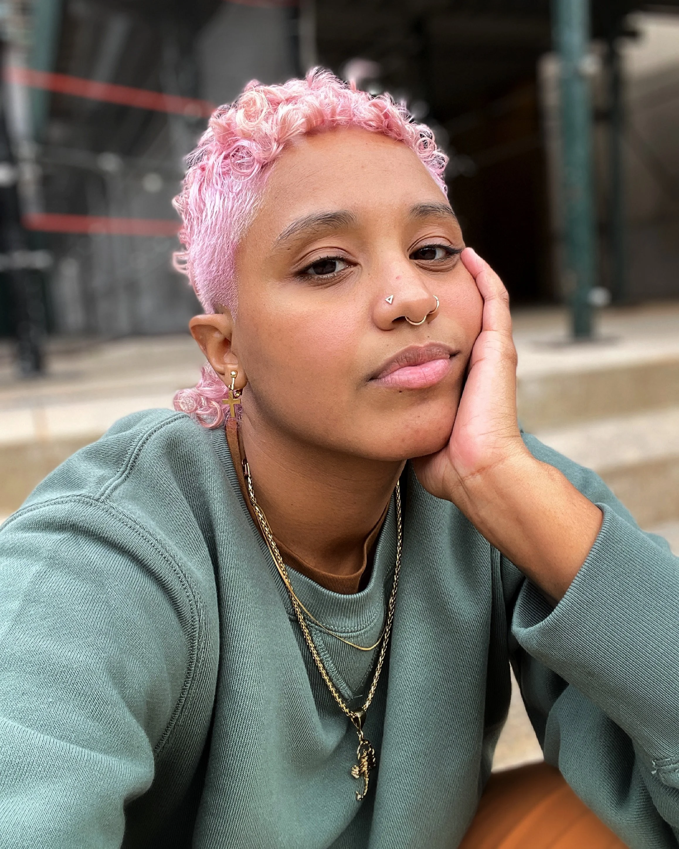 Ki is sitting down on a step with their left hand on their face. They have pink hair, a pastel green crew neck sweatshirt, a gold scorpion chain and and are looking direc