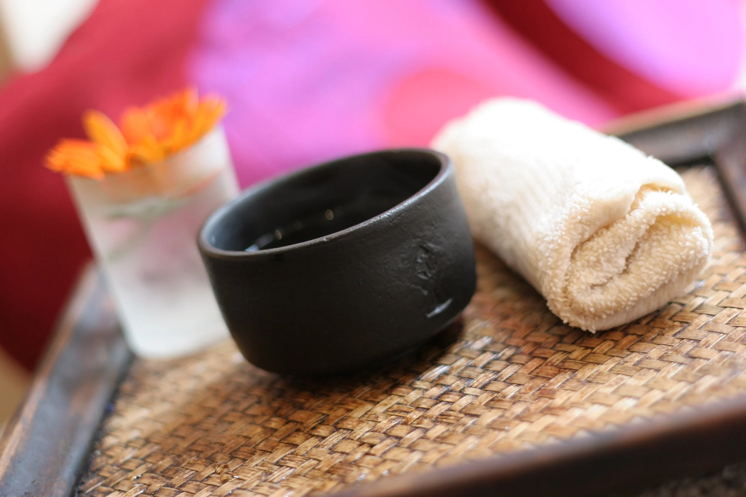 A black bowl and a rolled white towel on a wicker tray, with a small glass vase containing orange flowers in the background.