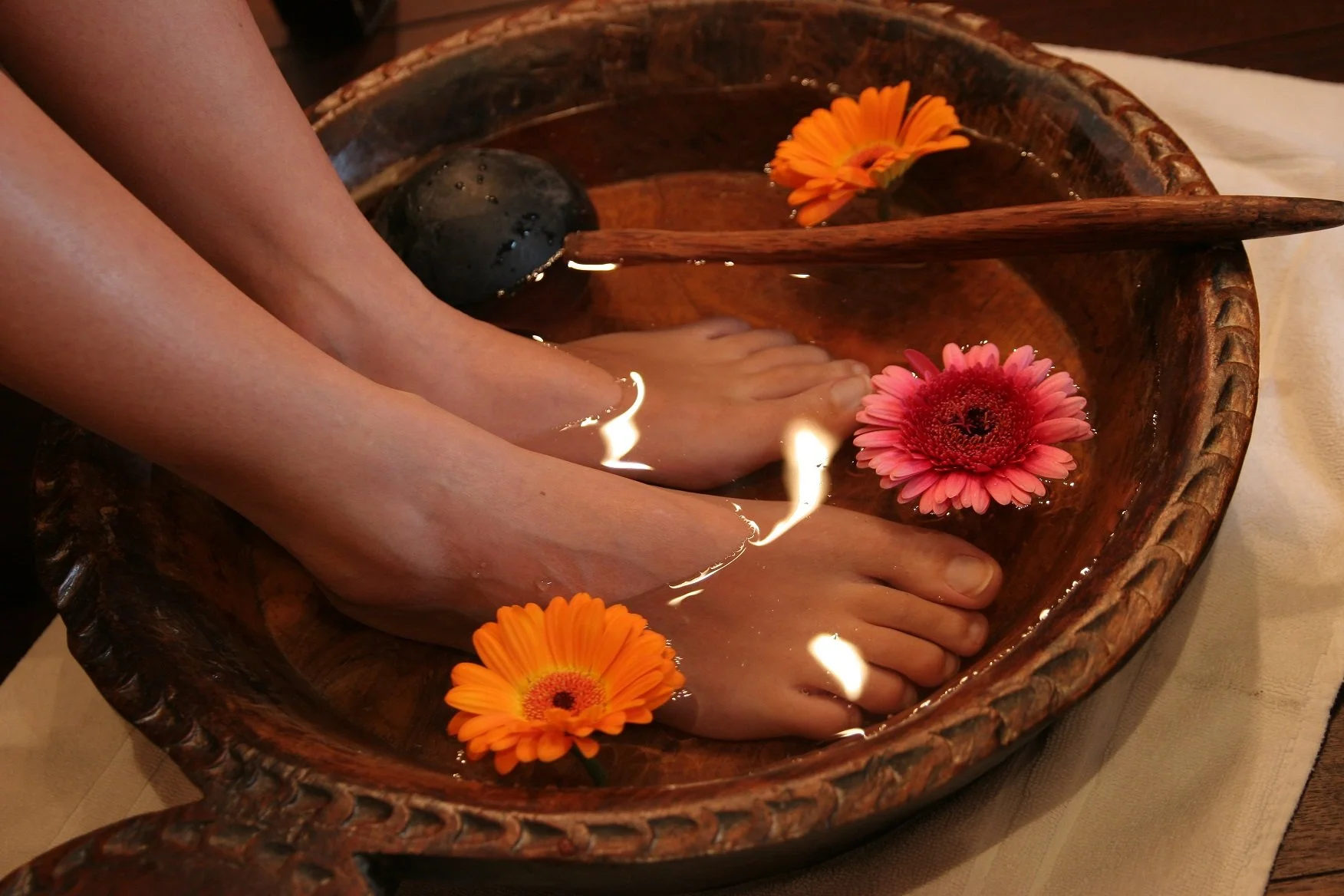 Feet soaking in a wooden bowl with orange and pink flowers floating on water.