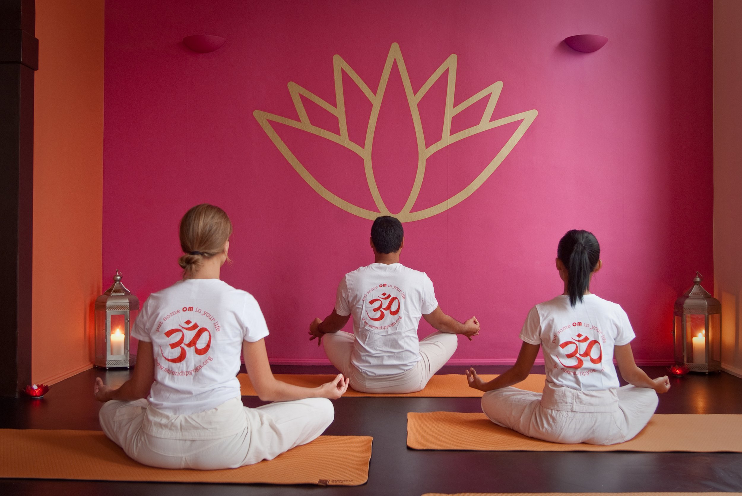 Three people practicing yoga in a studio with pink walls, lotus flower design on the wall, and candle lanterns on the floor.