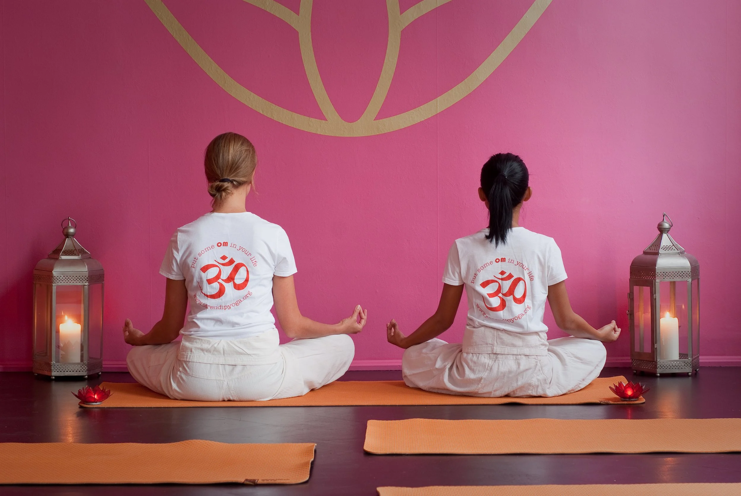 Two women practicing yoga in a pink room with candles and lanterns, sitting cross-legged with hands in a mudra position. They are wearing white t-shirts with a red Om symbol.
