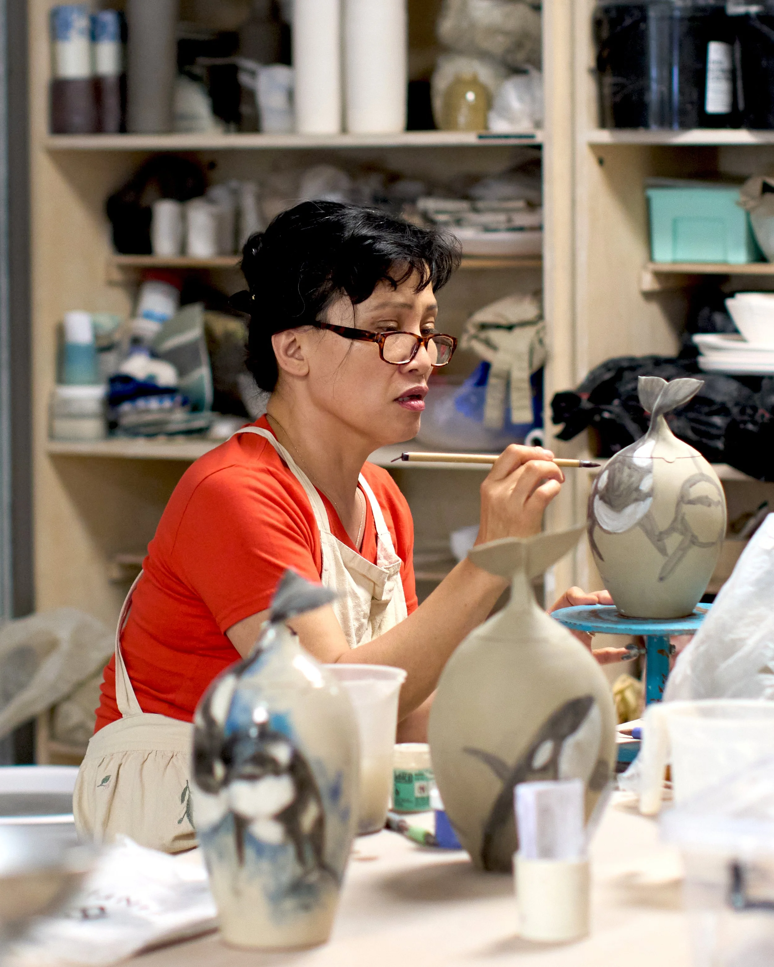 Person wearing glasses and a red shirt working on pottery in an art studio, surrounded by ceramic pieces.