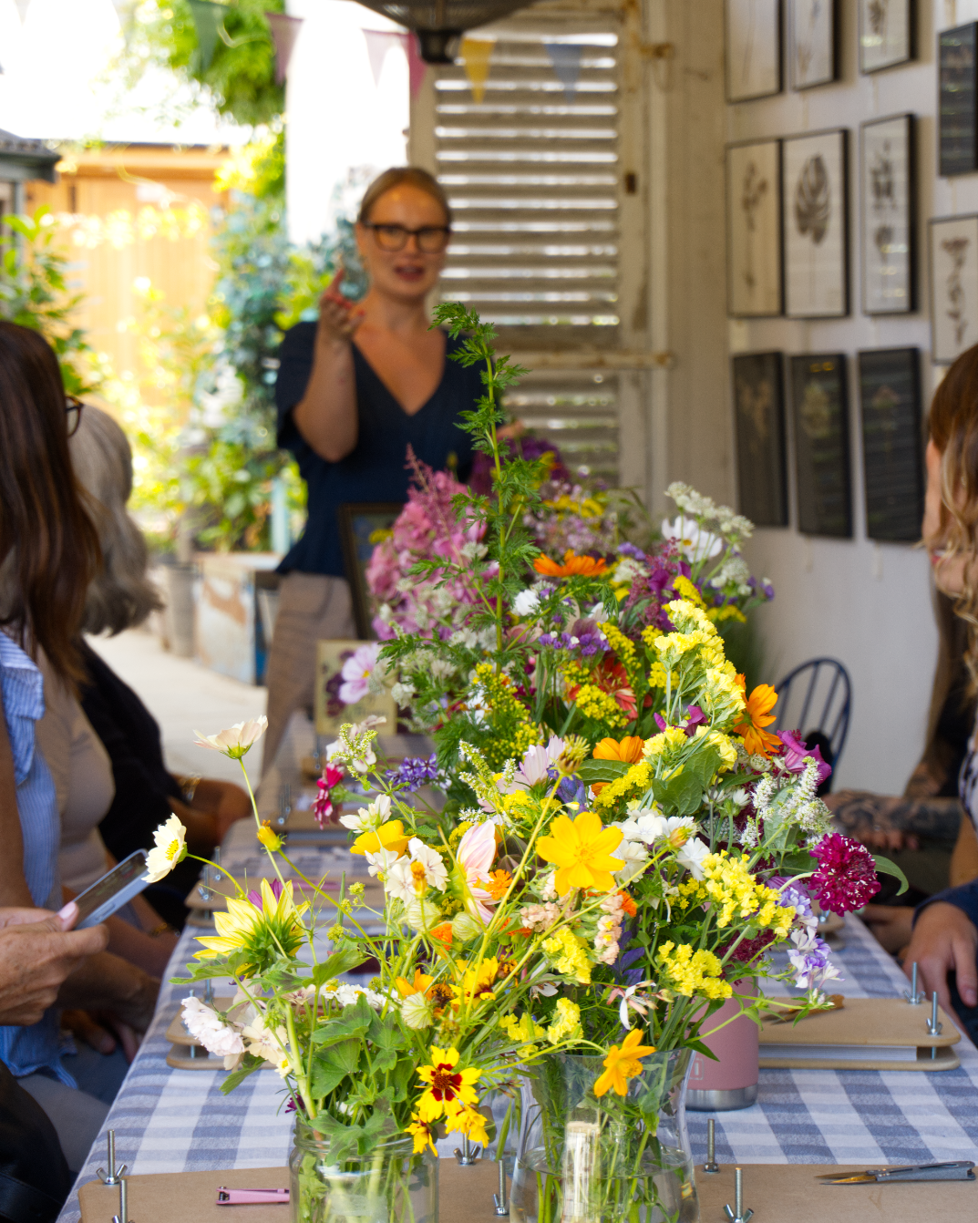 A woman standing and talking to a group of people seated around a table with a large colorful floral arrangement, inside a room with wall art and natural light.