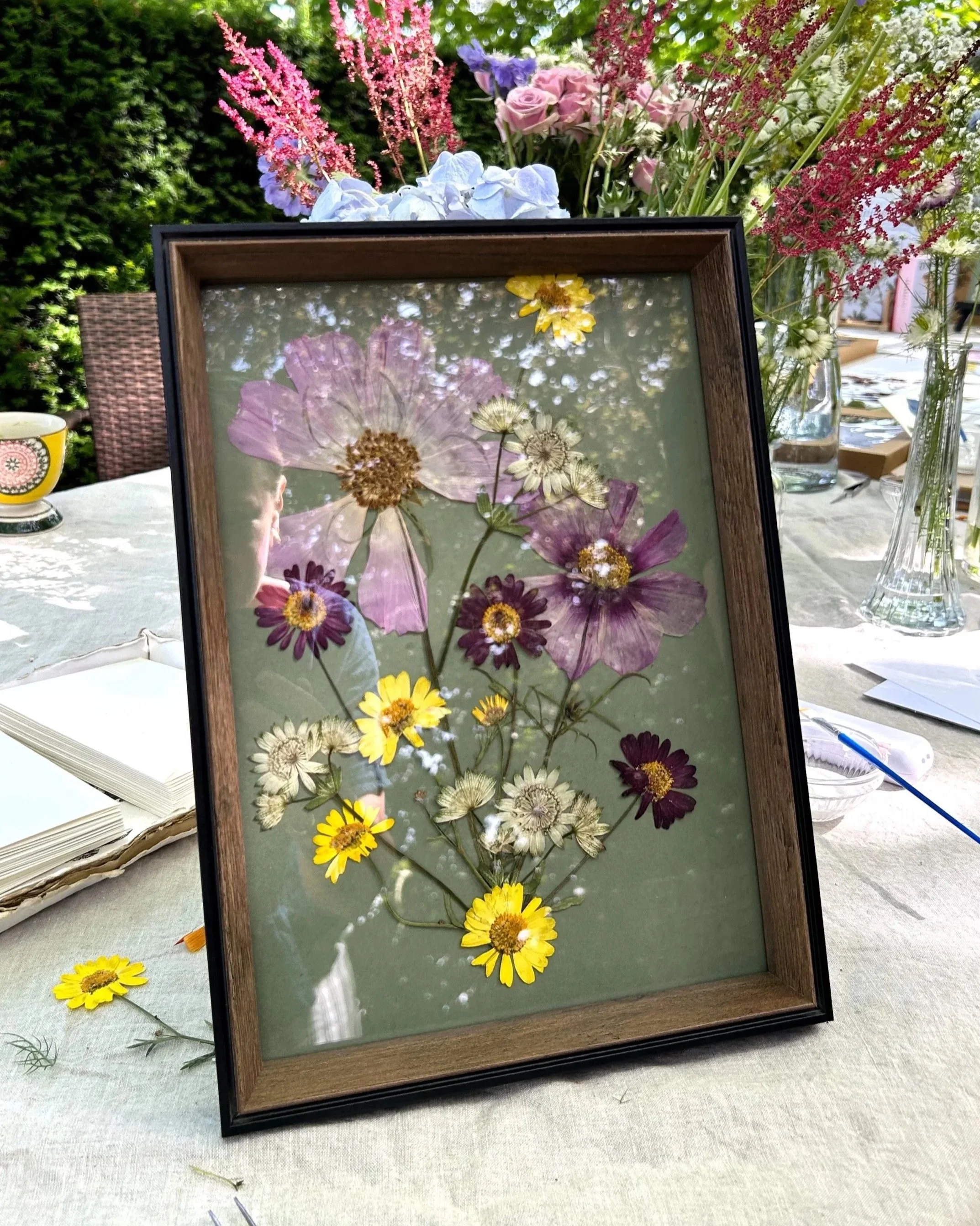 A framed collage of pressed flowers on a glass surface, with real flowers in vases and a table setting in the background at an outdoor gathering.