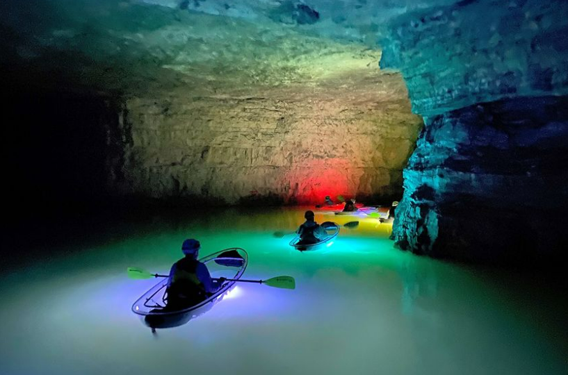 Group of people kayaking in illuminated cave with colorful lights in clear kayaks