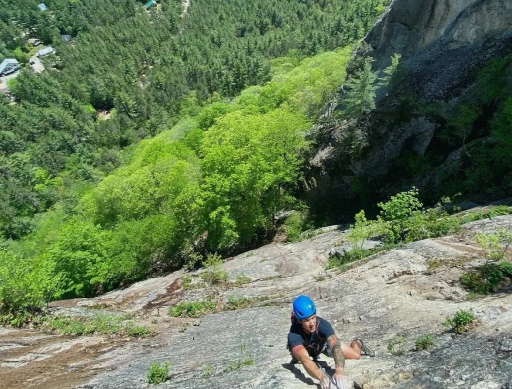 Climber in blue helmet scaling a steep rock face with lush green forest below.