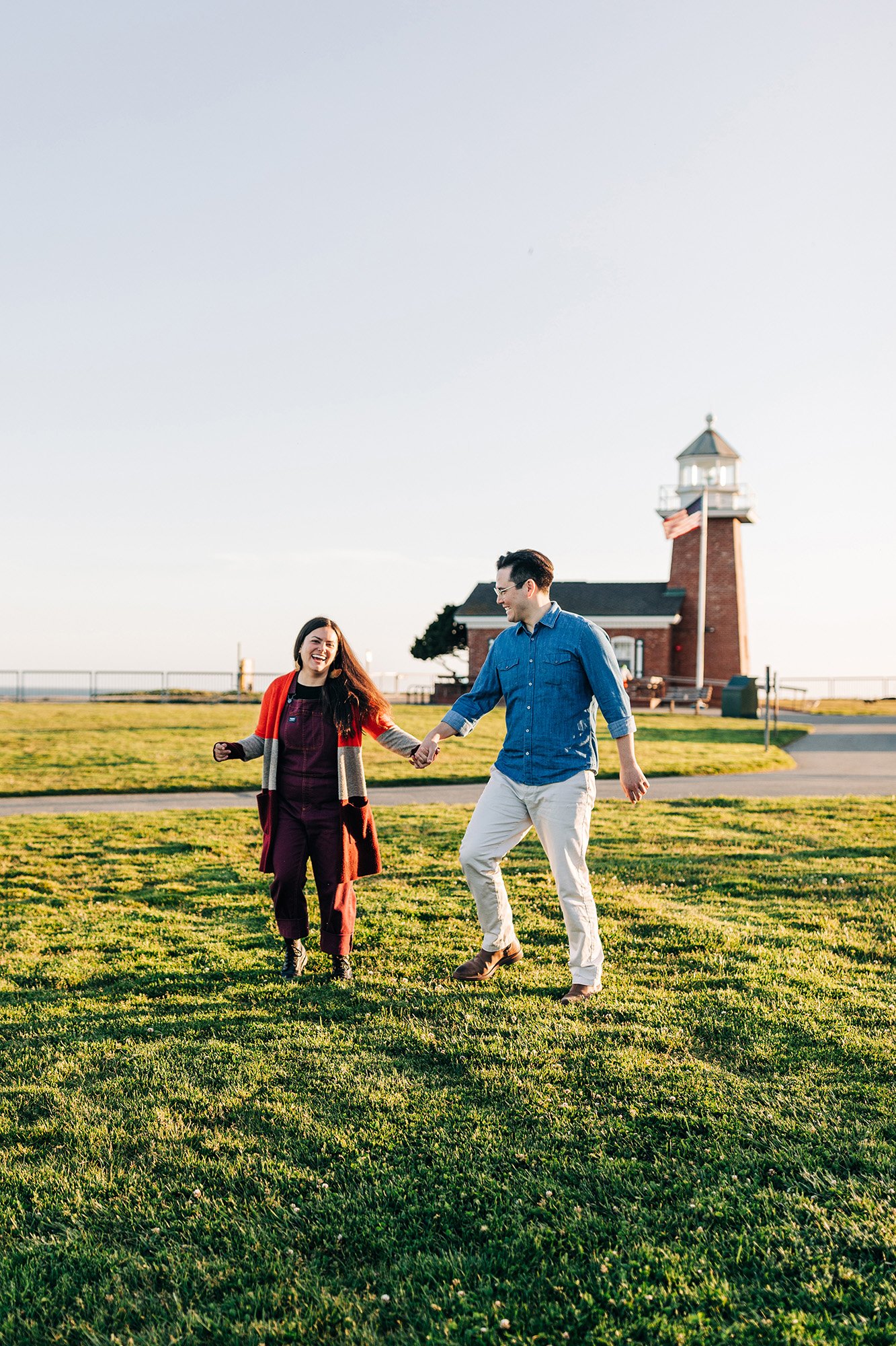 Chloe and Adam walk together on the greenery in front of a lighthouse in Santa Cruz, California.