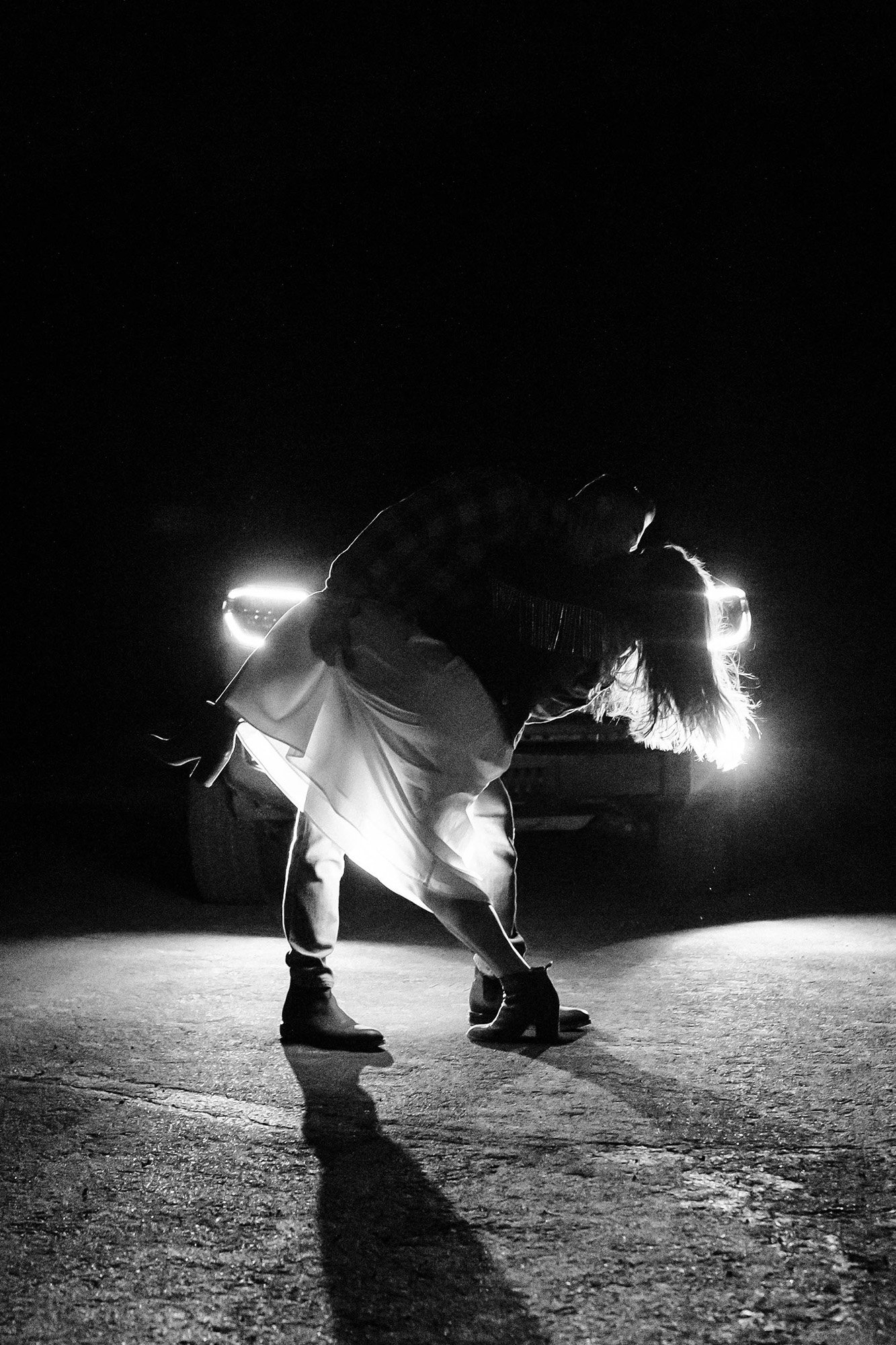 A couple shares a dramatic kiss in front of the headlights of a truck.