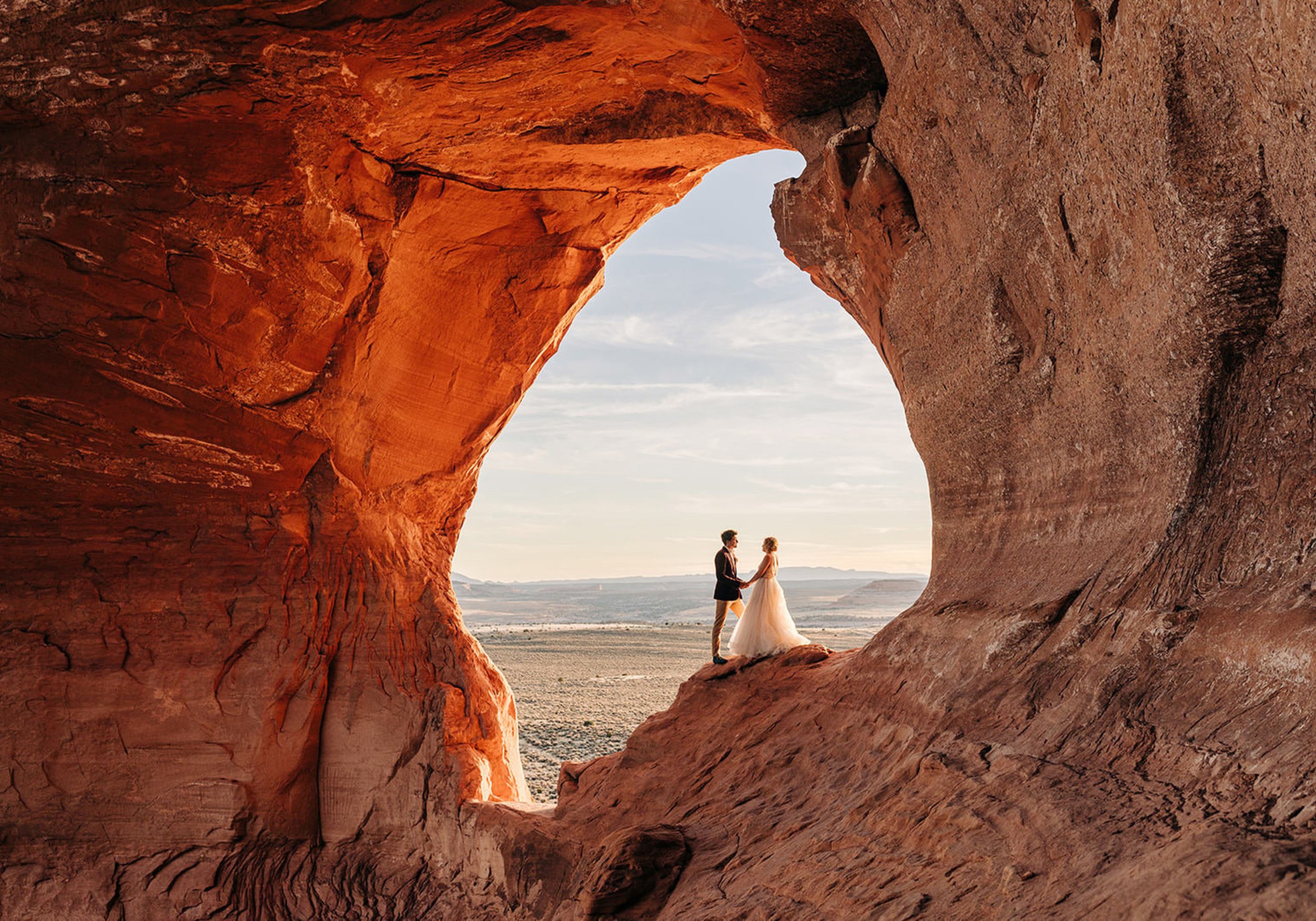 A wedding couple standing in the middle of a large rock arch. 