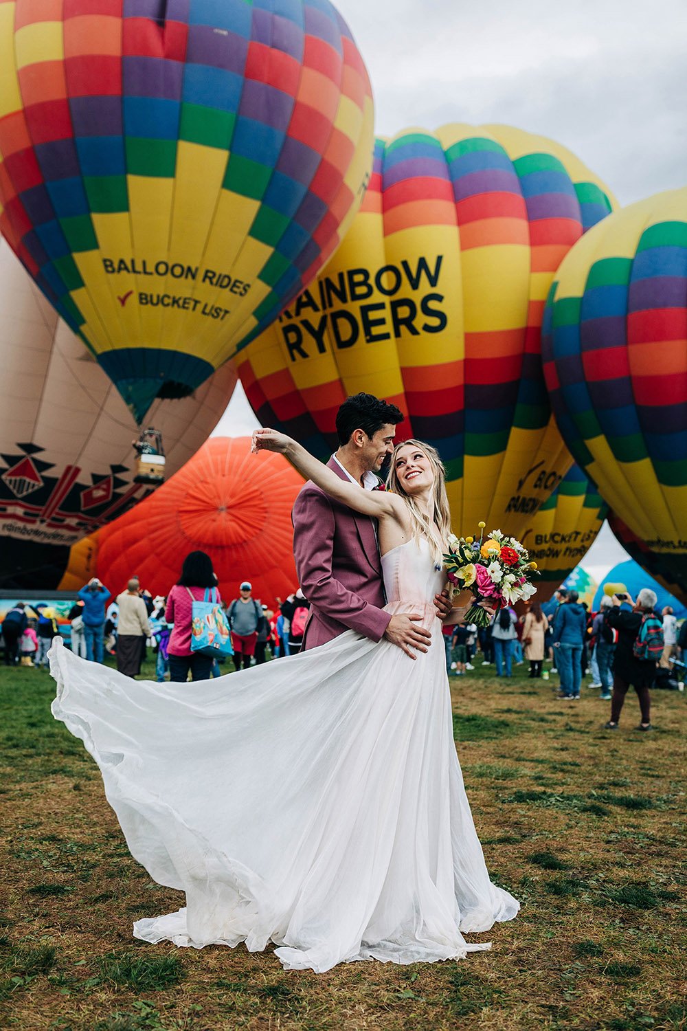 A woman tosses the end of her wedding dress while her husband holds her in front of hot air balloons.