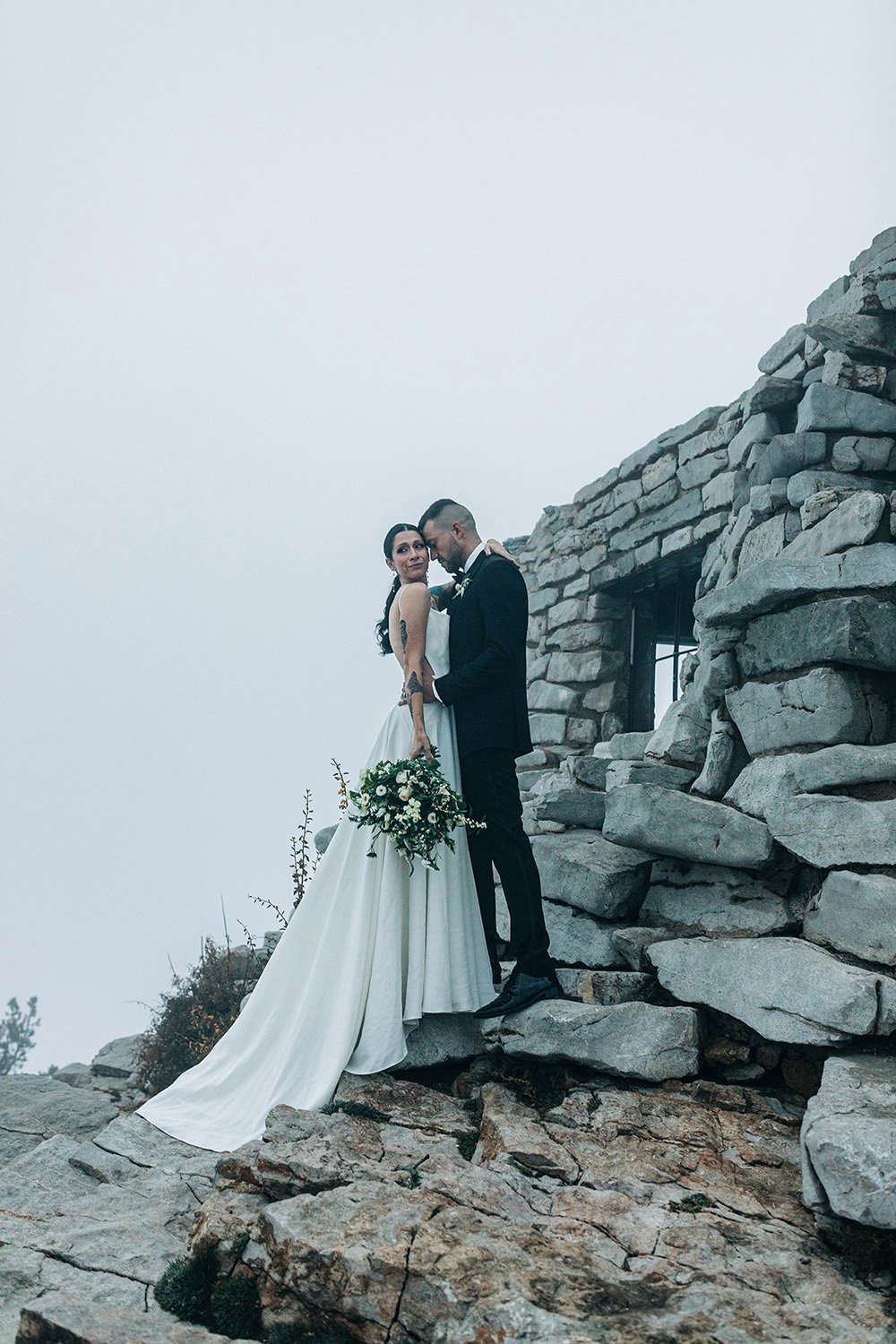 A couple cuddles on their wedding day by some ruins and a foggy view. 