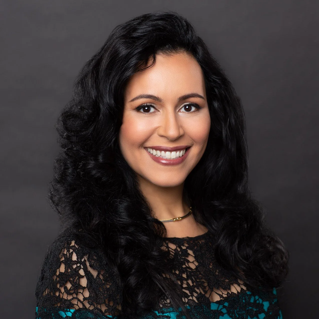 Woman with dark brown hair and radiant smile wearing a knit patterned top over a teal camisole