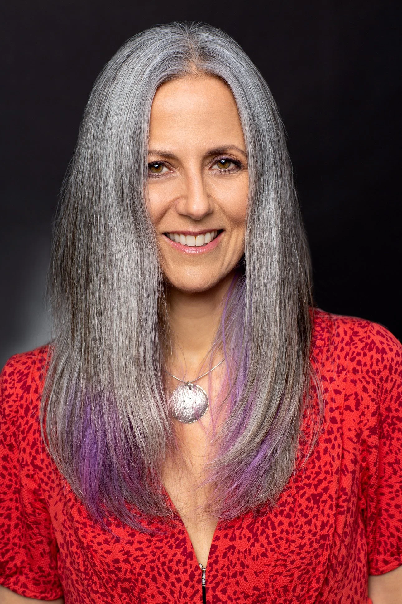 Headshot of woman with naturally grey hair wearing a red patterned wardrobe