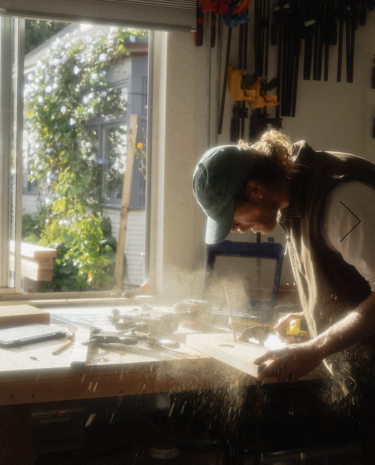 A person wearing a green cap working on a woodworking project at a cluttered workbench inside a workshop, with sunlight coming through a window and greenery visible outside.