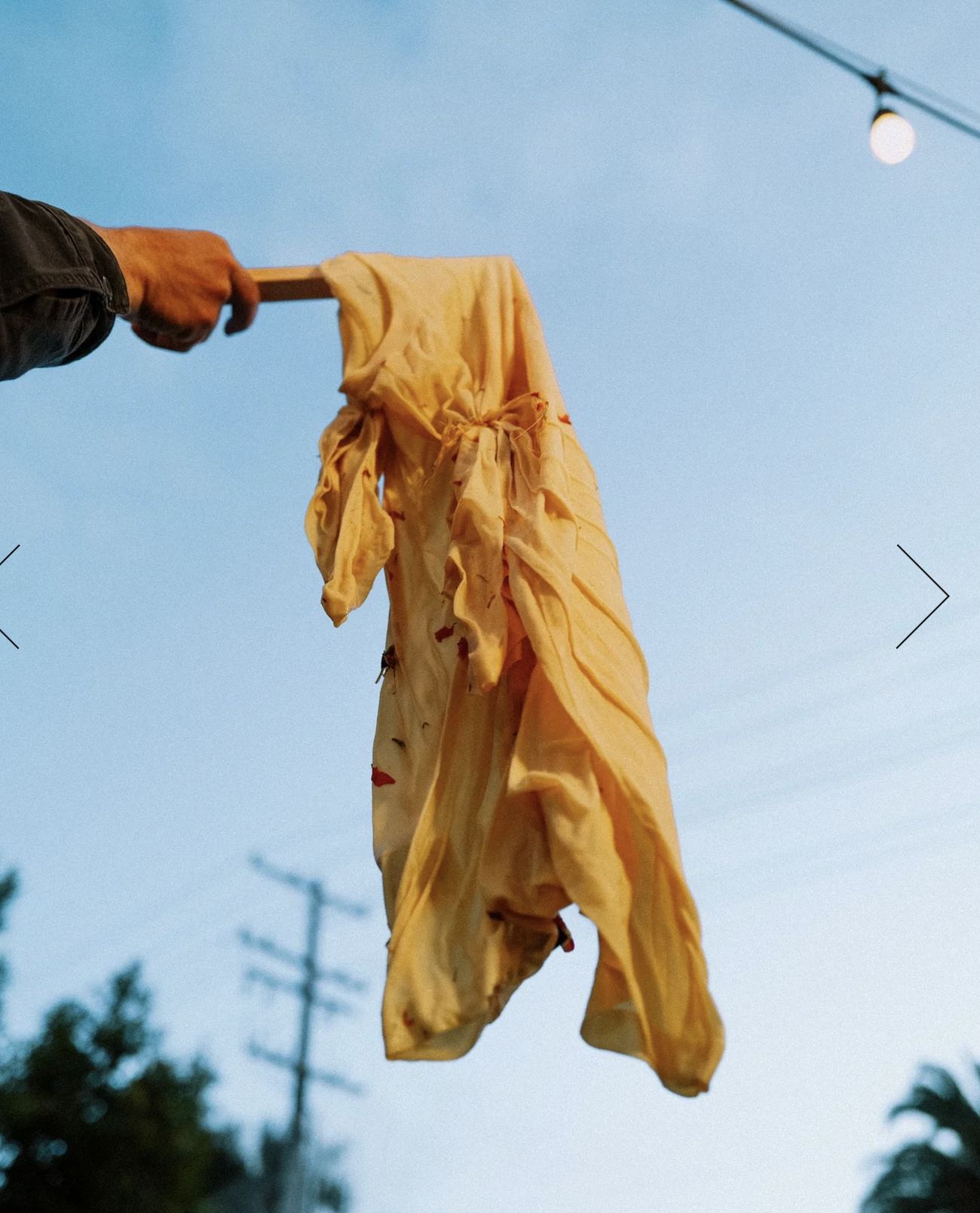 Yellow fabric hanging on a wooden stick held by a person's hand, against a clear sky with a string of outdoor lights and trees visible in the background.