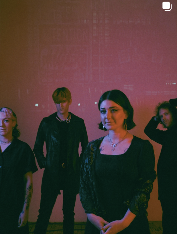 Group of four people standing against a pinkish-red wall with a projected cityscape in the background, three women and one man, dressed in black, posing for a photo.