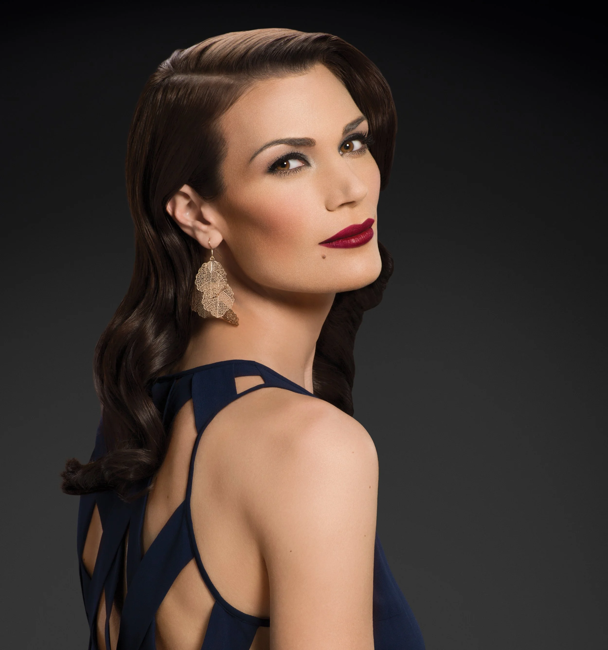 Woman in blue dress with elegant hairstyle and gold leaf earrings, posing against a dark background.