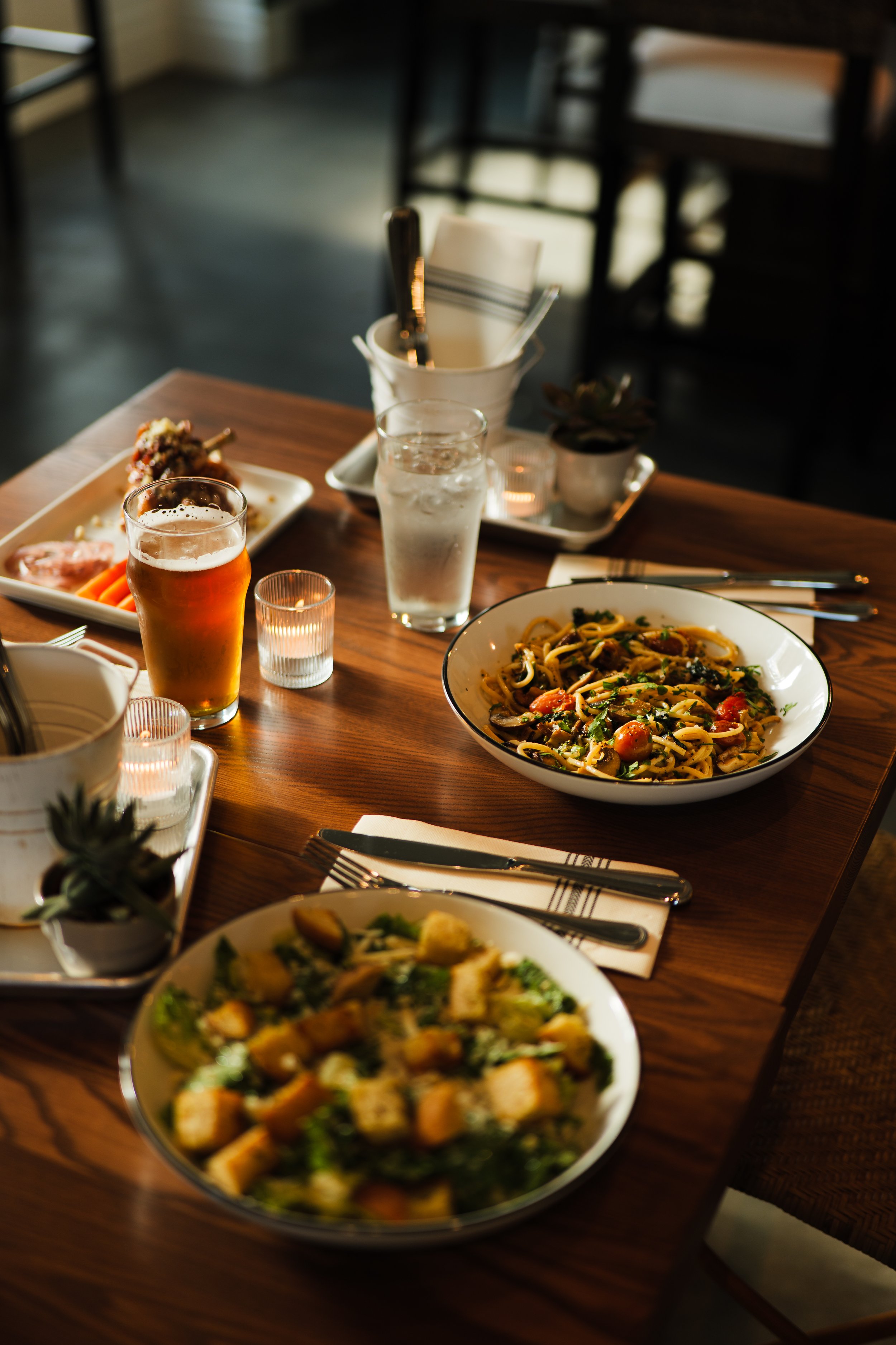 Restaurant photography featuring a fully plated table of pasta, a glass of beer, a glass of water, candles, and small potted plants in a cozy cafe.