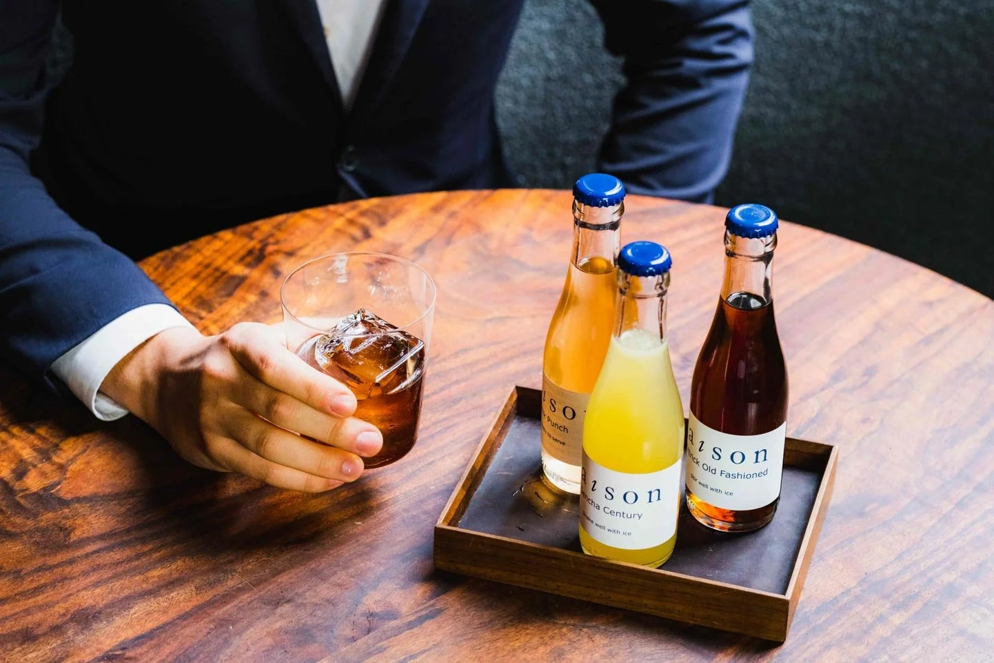 Person in a suit holding a glass of iced cola, sitting at a wooden table with a tray of three small bottles of Japanese soda.