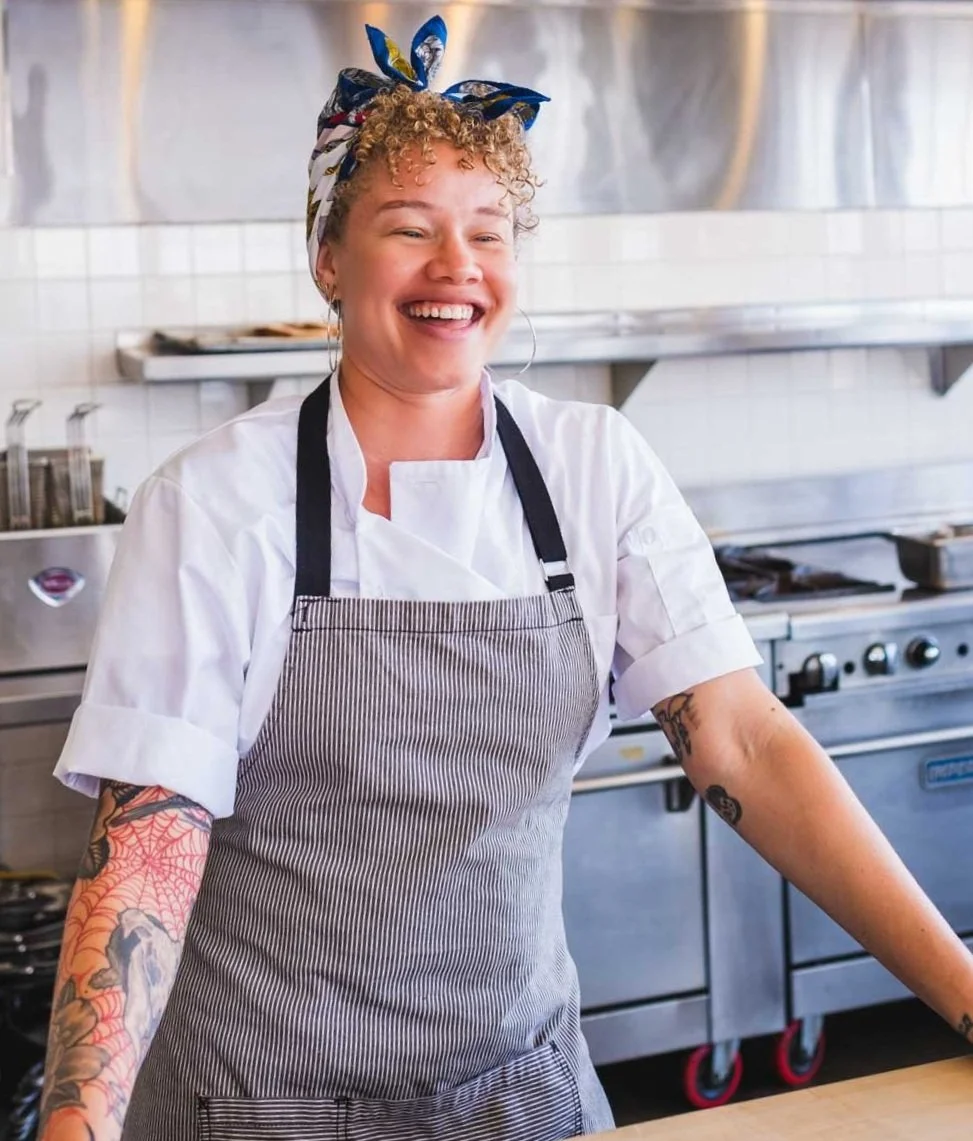 A smiling woman with tattoos and curly hair, wearing a headscarf, white chef's coat, and a striped apron, standing in a commercial kitchen.