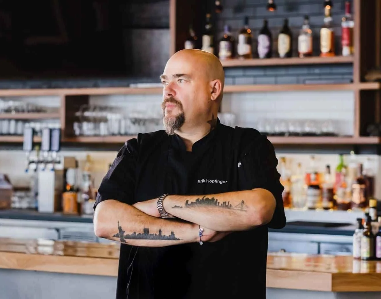 A male chef with crossed arms standing in a restaurant kitchen, with no visible smile. Tattoos of horizon skyline and Brooklyn Bridge on his forearms, wearing a black chef's uniform with his name 'Erik Hopfinger' visible. Background includes shelves 