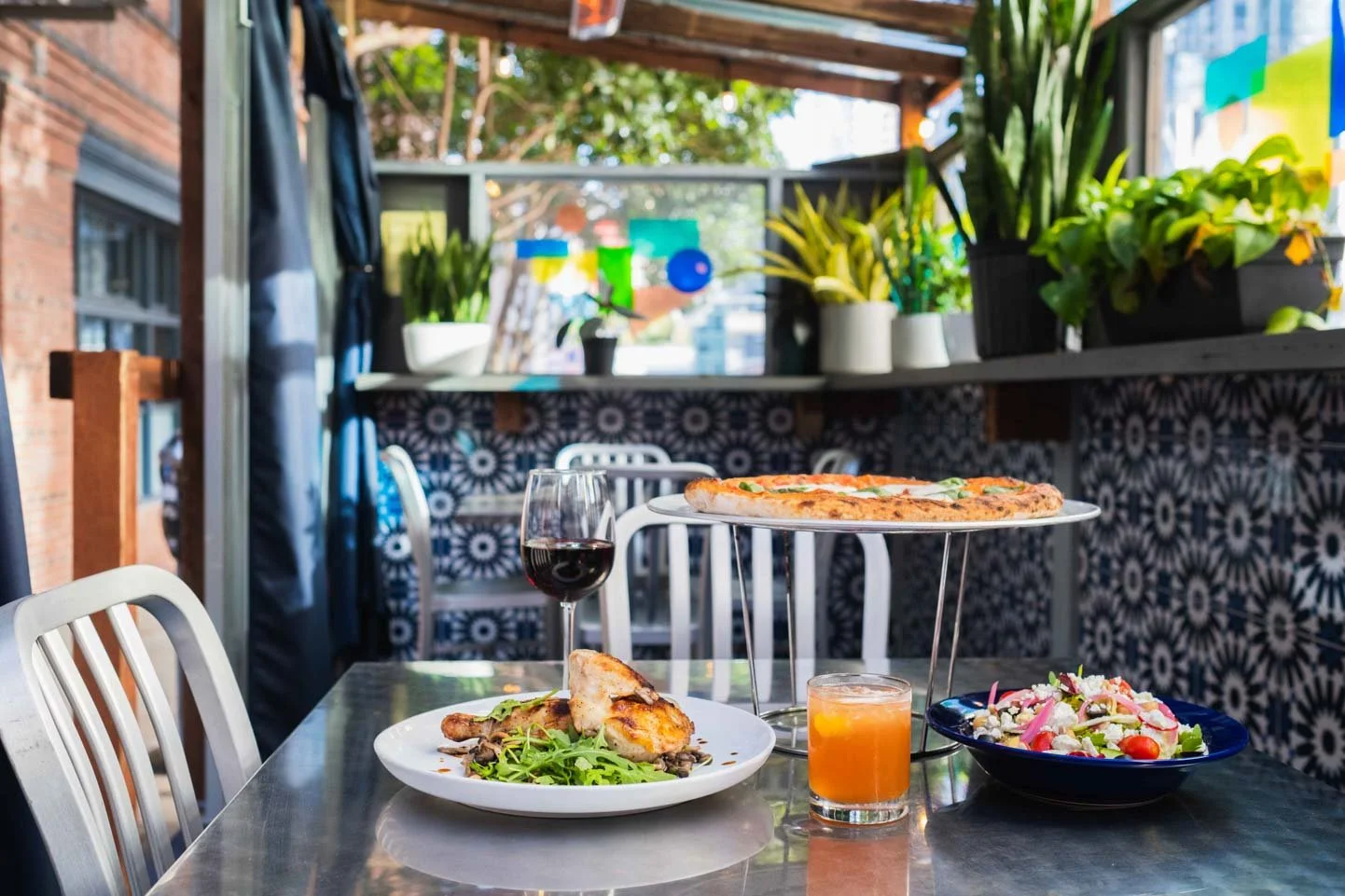 Outdoor dining table with pizza, wine, salad, and a glass of orange drink, set on a patio decorated with potted plants and colorful tiles.