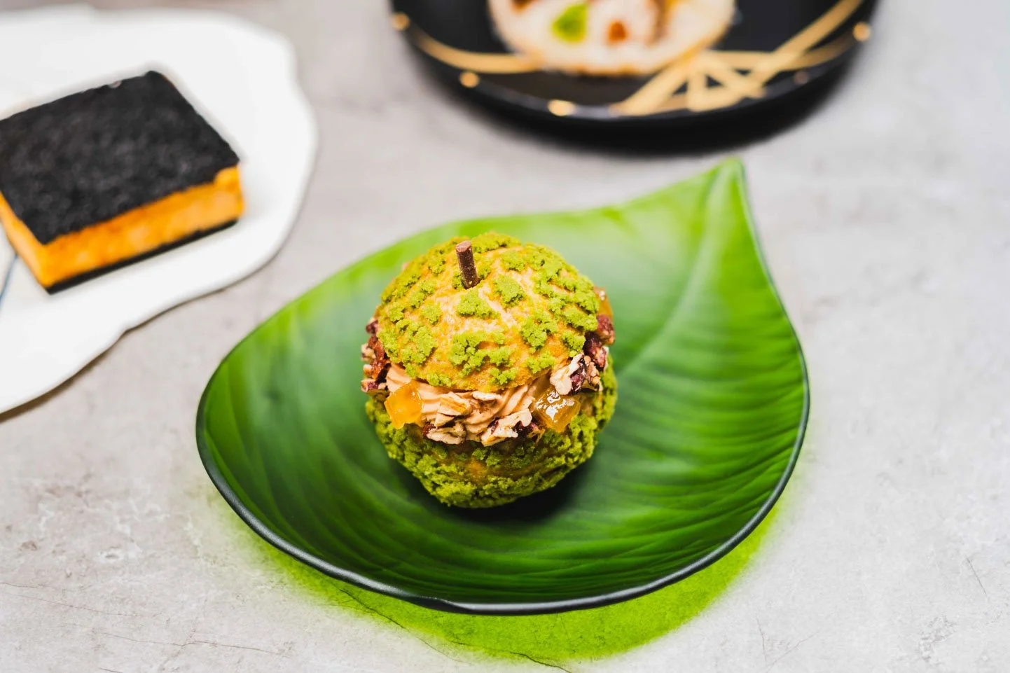 A green leaf-shaped plate holding a scoop of green ice cream with a layer of whipped cream and chopped nuts, garnished with a chocolate stick.