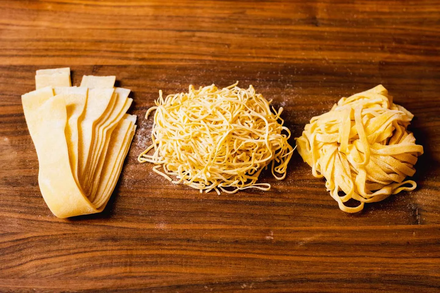 Three types of shredded pasta on a wooden surface, from left to right: lasagna sheets, shredded cheese, and fettuccine pasta.