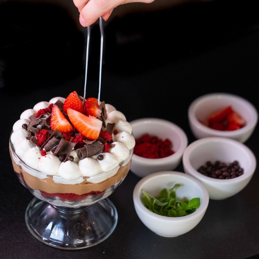 A layered dessert in a glass bowl topped with strawberries, chocolate shavings, and cream, with a hand placing tongs on top. Small bowls with various toppings like strawberries, chocolate chips, and green garnish are visible on a black surface.