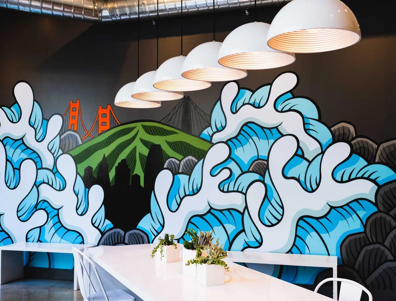 A colorful mural featuring waves, a mountain, and a bridge in an indoor setting with white tables and hanging white pendant lights.