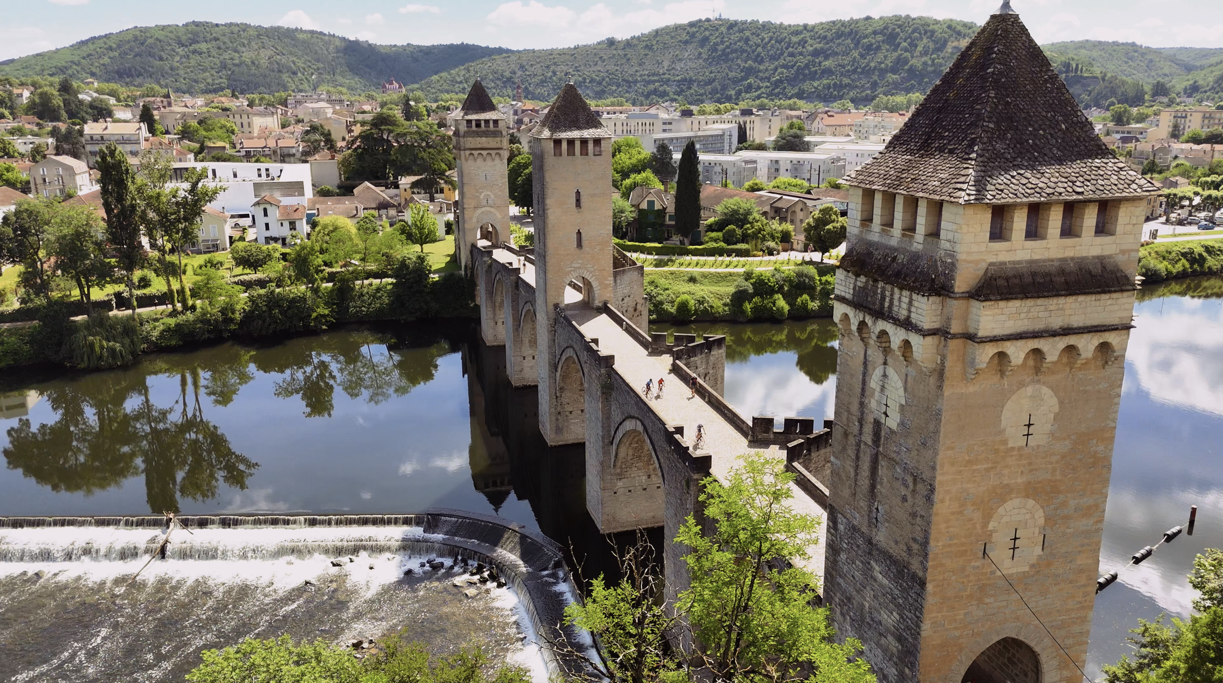 A scenic view of a historic bridge with towers crossing over a river, with modern buildings and green hills in the background.