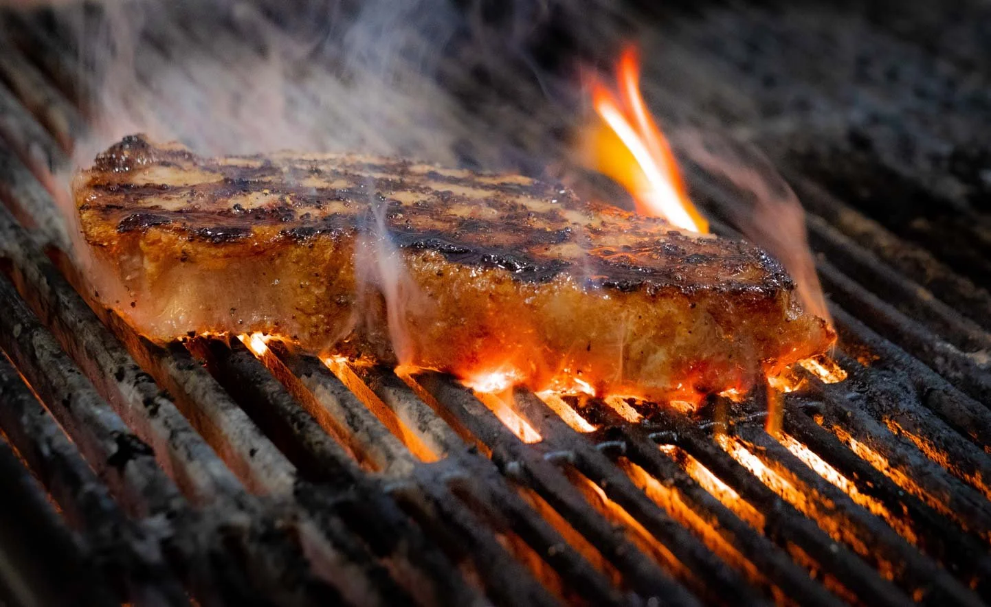 A close-up of a steak grilling on a barbecue, with flames and smoke visible.